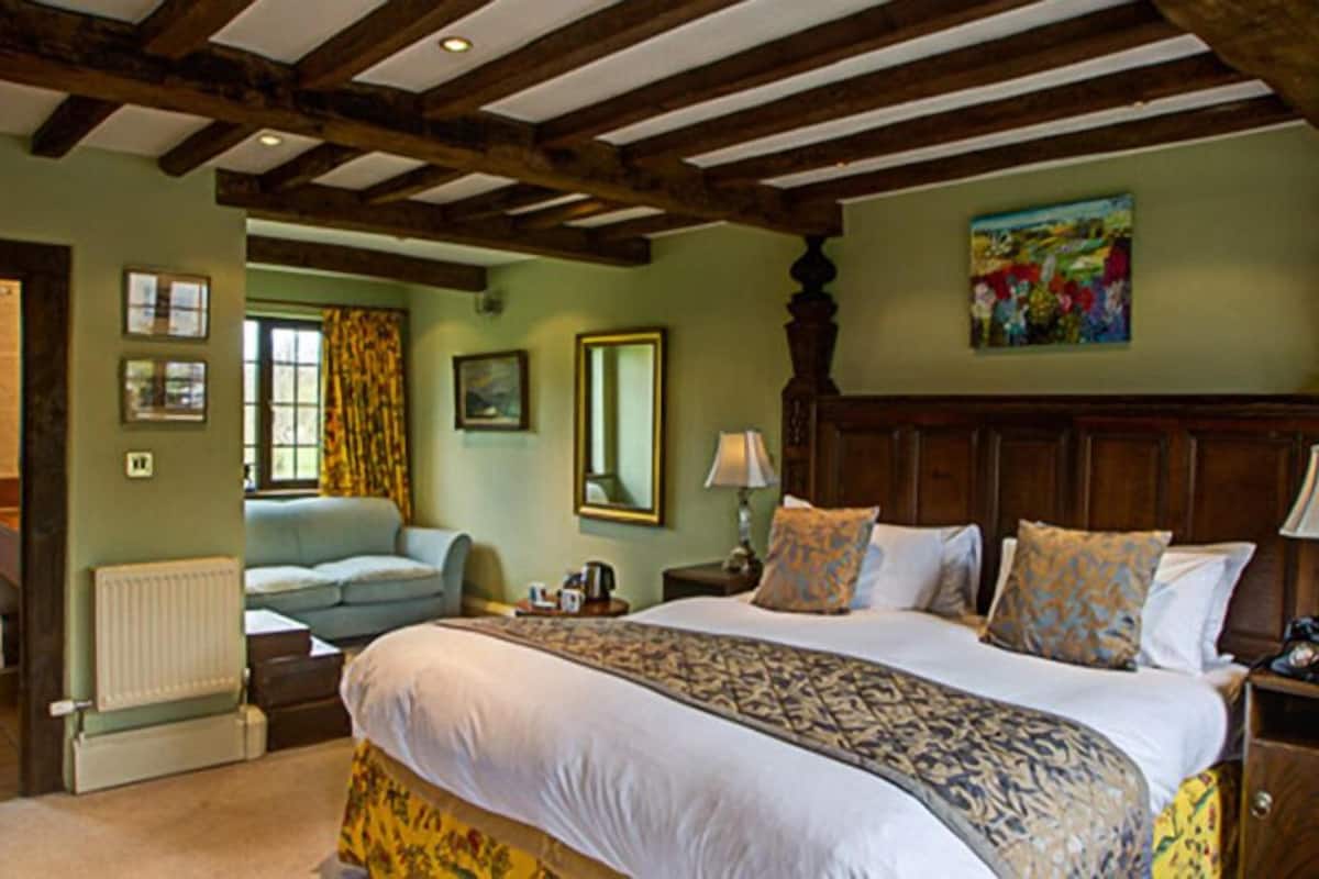 A bedroom with a large bed, wooden ceiling beams, a radiator, a window with curtains, a painting on the wall, and a sofa in the adjacent room.