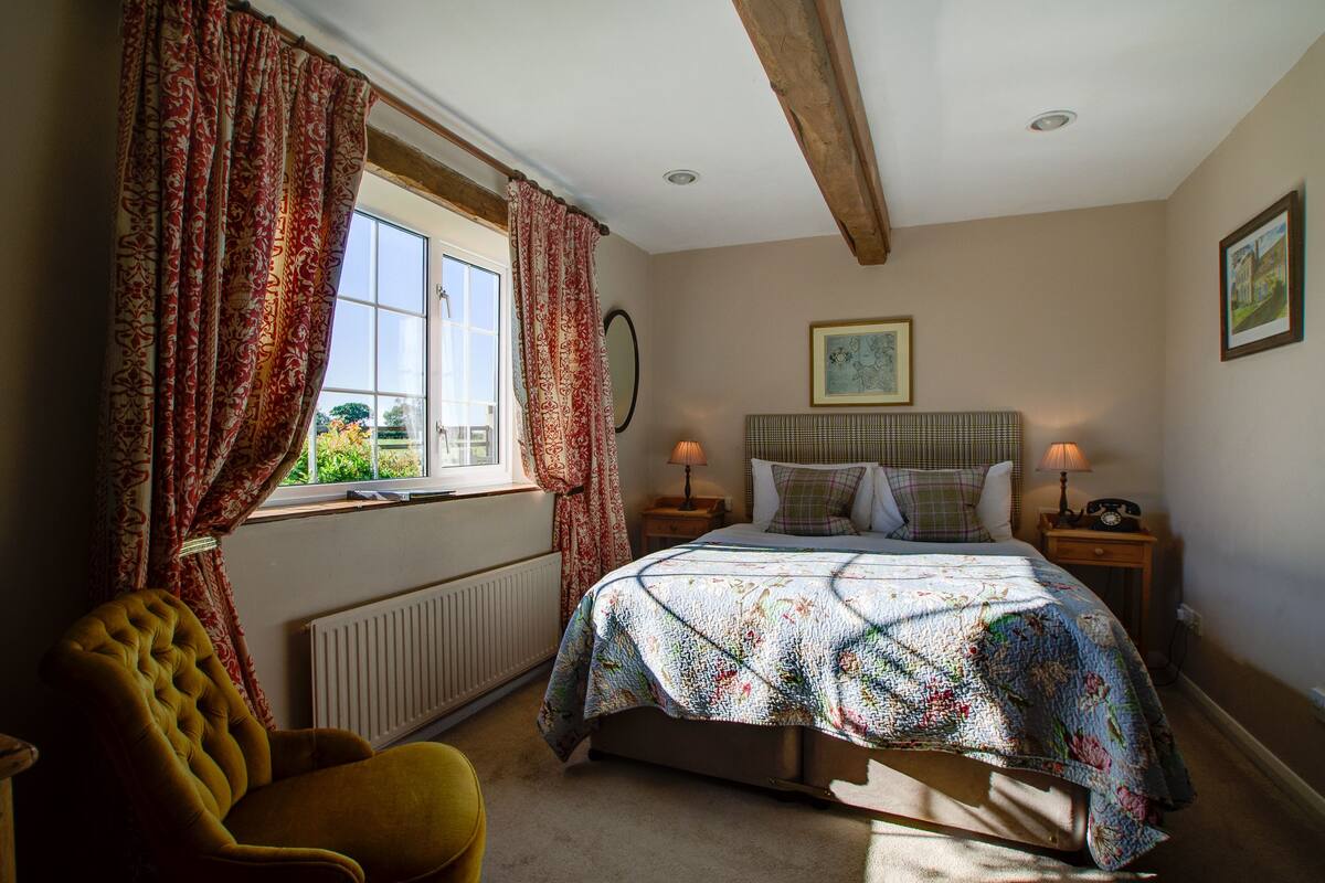 A bedroom with a bed, two bedside tables with lamps, a chair, a window with curtains, and a radiator.