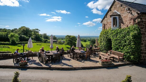 Un café en plein air avec des tables et des chaises en pierre, entouré d’une végétation luxuriante et d’un bâtiment en pierre.