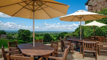 An outdoor seating area with wooden tables and chairs, two large umbrellas, and a view of the countryside.
