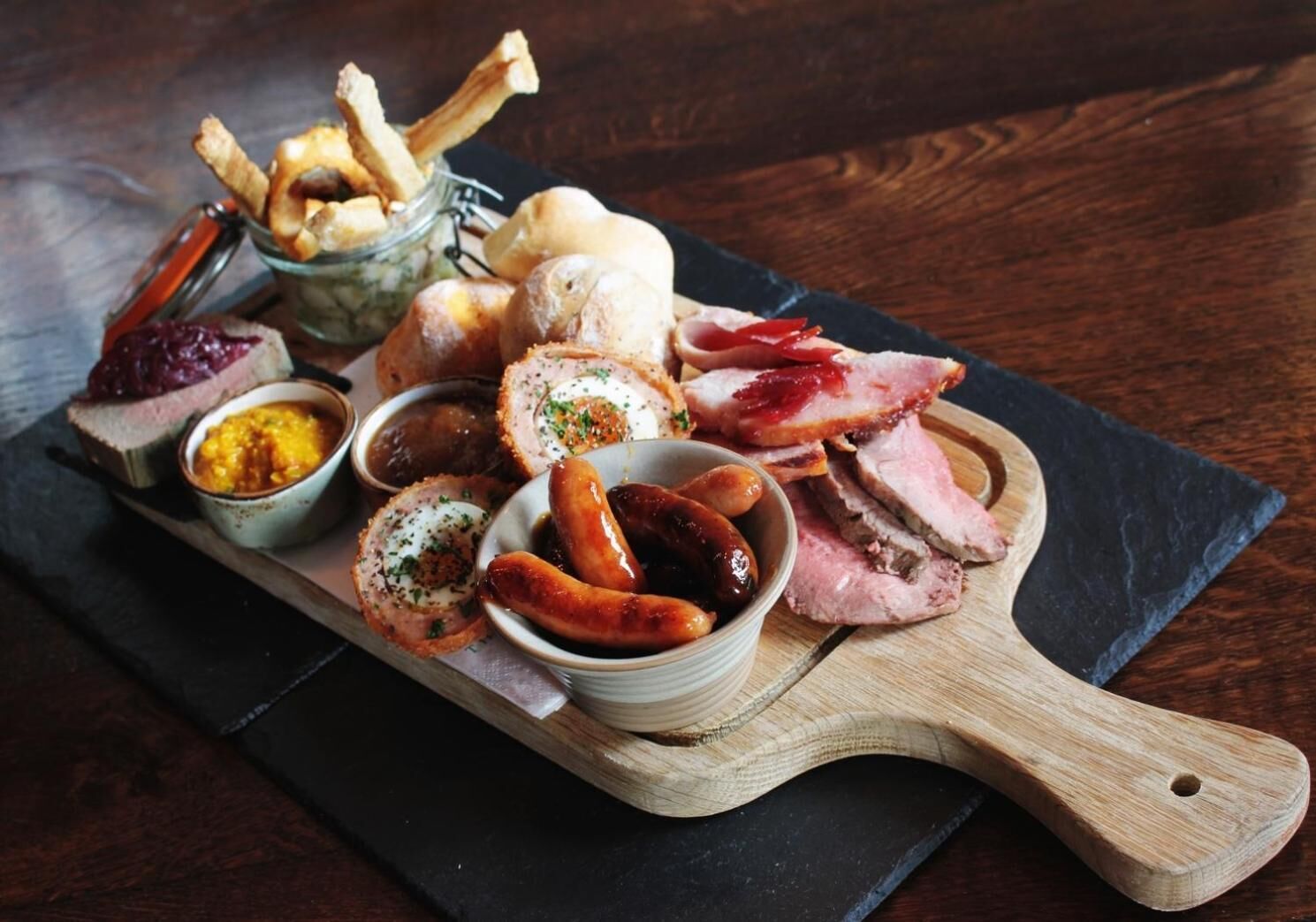 A wooden board with various food items including sausages, bread, and a pastry.