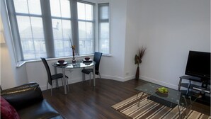 Una sala moderna con un sofá de cuero, una mesa de comedor de vidrio con sillas, un televisor de pantalla plana y un ventanal con persianas.