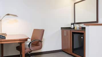 A hotel room with a wooden desk, a brown leather chair, a desk lamp, a refrigerator, and a coffee maker.