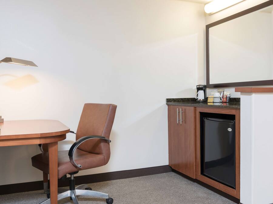 Habitación de hotel con un escritorio de madera, una silla de cuero marrón, una lámpara de escritorio, un refrigerador y una cafetera.