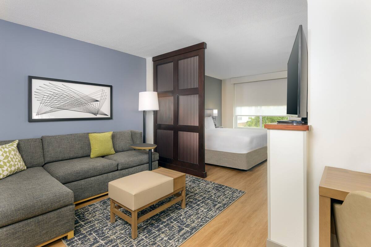Quarto de hotel moderno com um sofá cinzento, uma pequena mesa de madeira, uma cama com edredom branco, uma TV fixada na parede e uma janela grande.