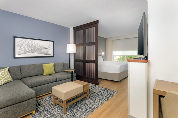Una habitación de hotel moderna con un sofá gris, una pequeña mesa de madera, una cama con edredón blanco, un televisor montado en la pared y un ventanal amplio.