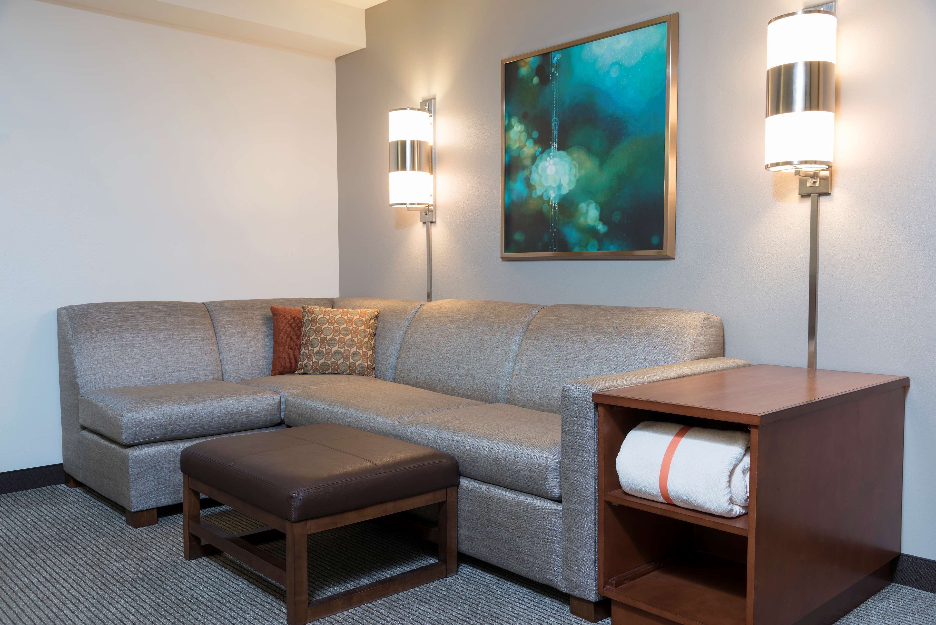 A modern hotel room with a grey sectional sofa, a wooden bench, a wooden side table with a rolled towel, and two wall-mounted lamps.