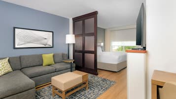 A modern hotel room with a grey sofa, a small wooden table, a bed, a TV, and a large window.
