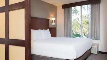 A hotel room with a large bed, a nightstand with a lamp, a window with curtains, and a view of greenery outside.
