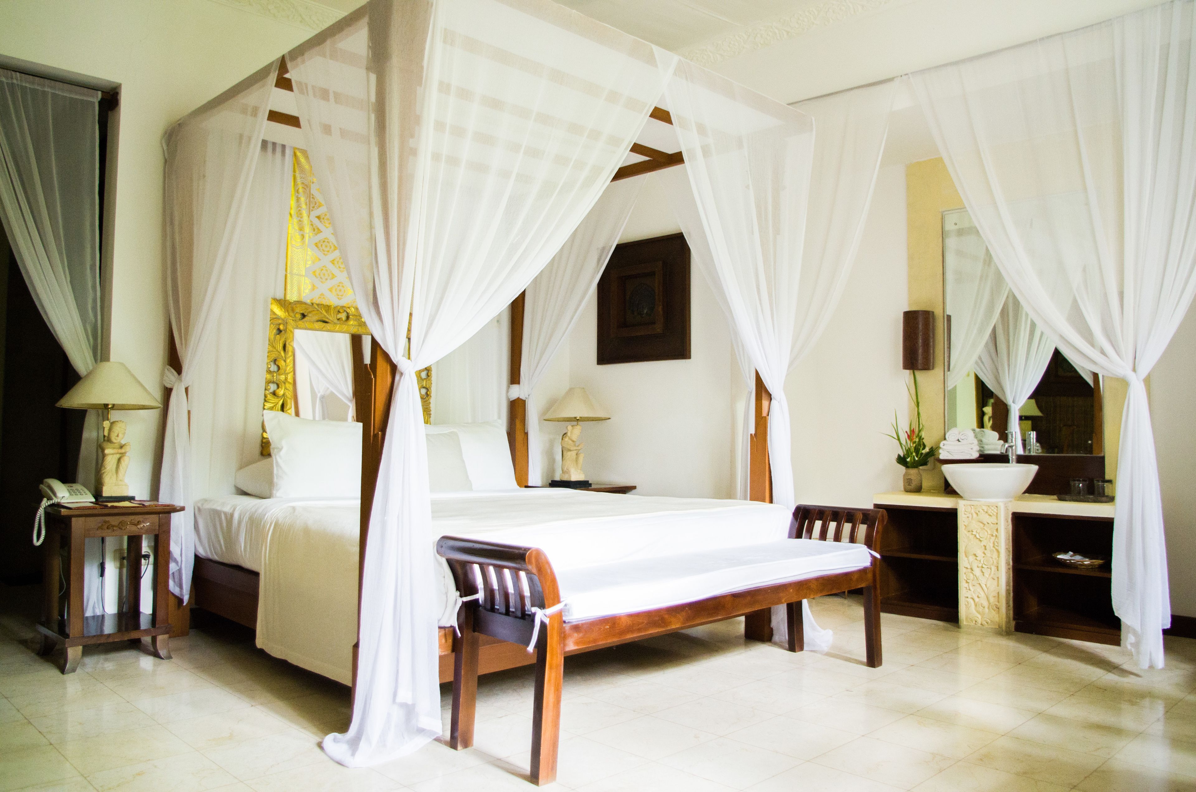 A four-poster bed with white drapes, a wooden bench, a bedside table with a lamp, and a mirror above the sink.