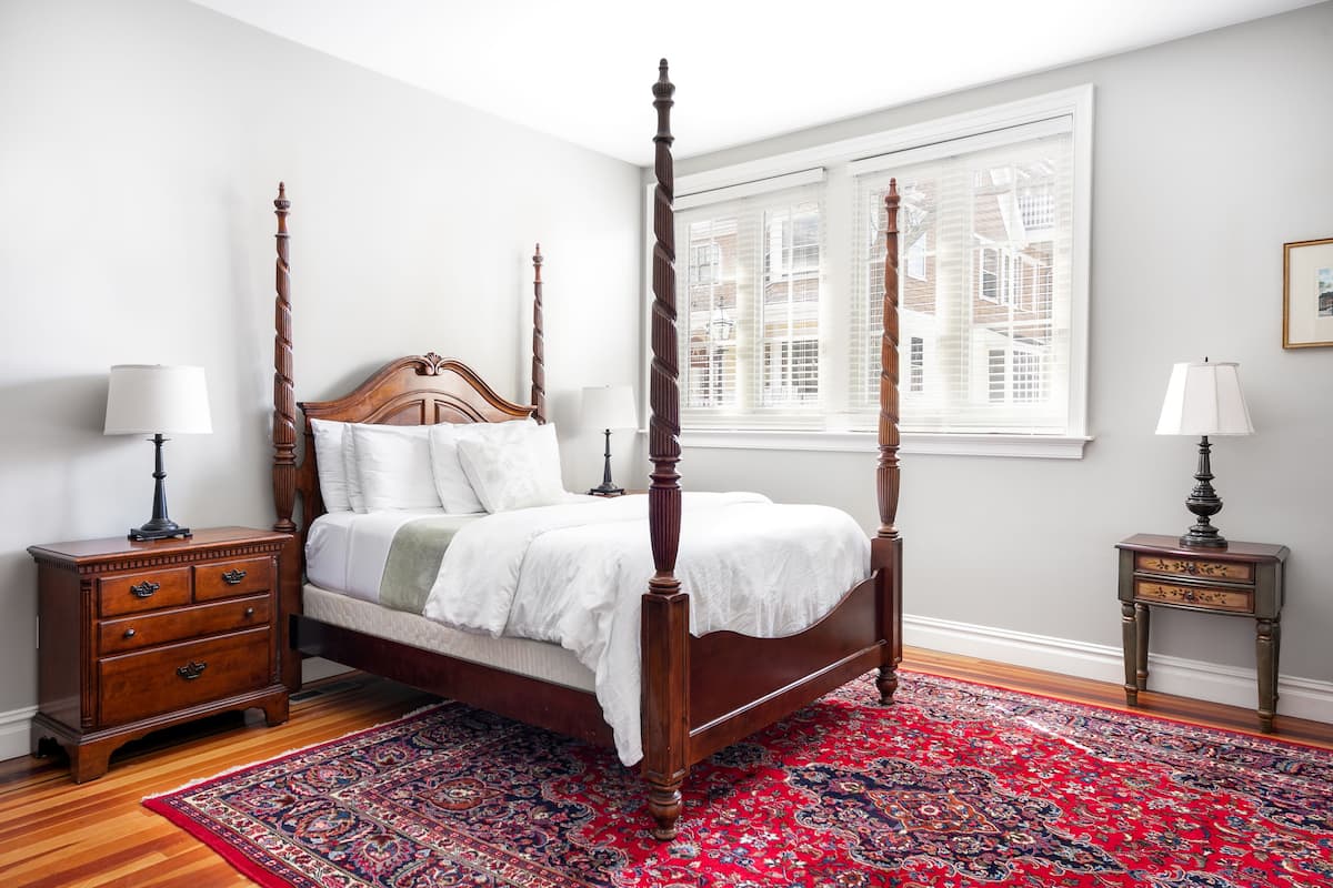 A four-poster bed with a wooden headboard, a red patterned rug, and two nightstands with lamps.