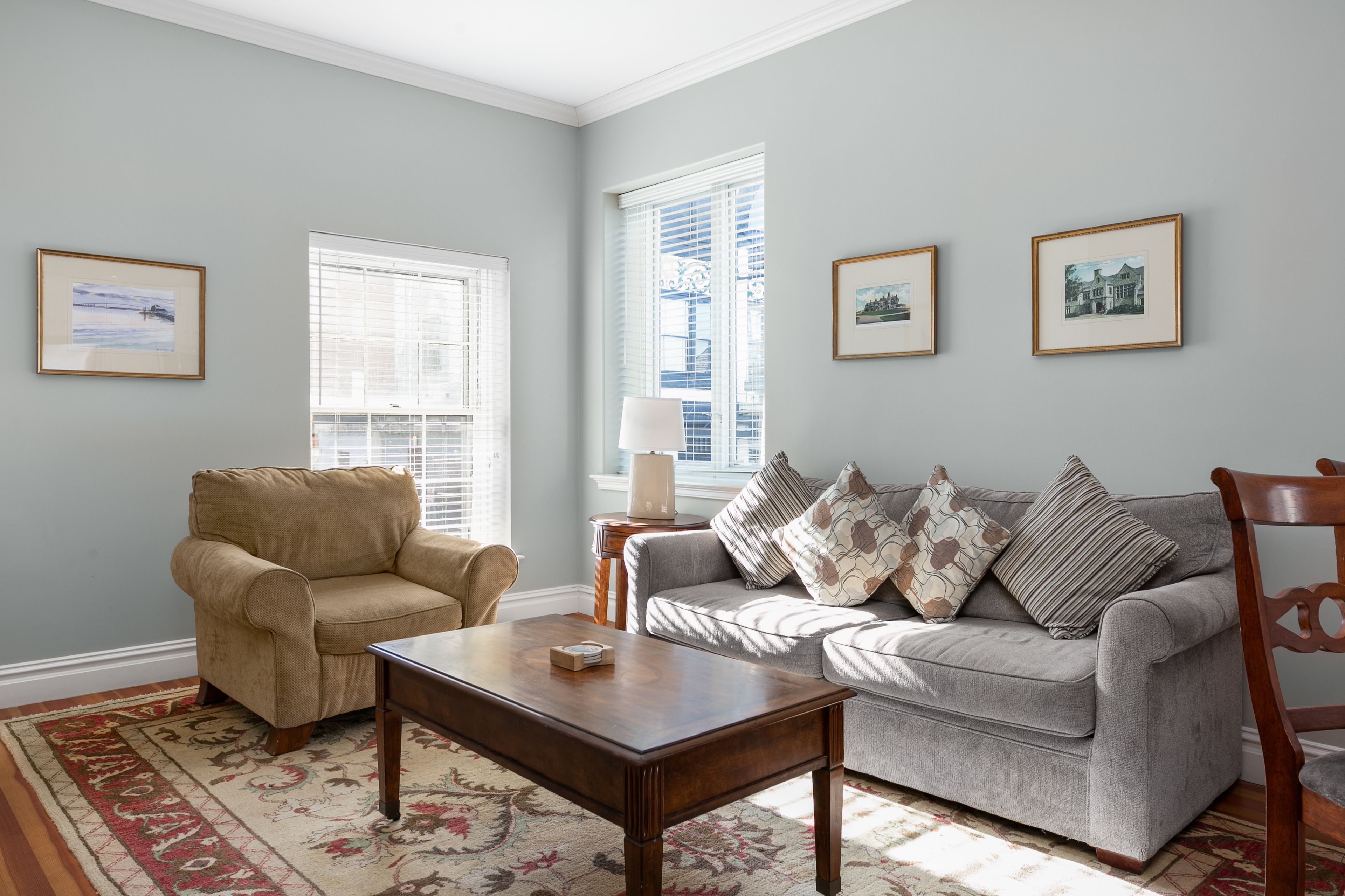 A living room with a beige armchair, a grey sofa with patterned cushions, a wooden coffee table, and a floral rug. There are framed pictures on the walls and a window with blinds.