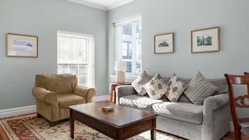 A living room with a beige armchair, a grey sofa with patterned cushions, a wooden coffee table, and a floral rug. There are framed pictures on the walls and a window with blinds.