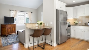 Una cocina con armarios blancos, un refrigerador de acero inoxidable y un comedor con dos sillas y una mesa.
