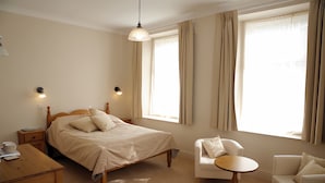 Un dormitorio con una cama, dos mesitas de noche, una silla, una mesita pequeña y una ventana con cortinas.