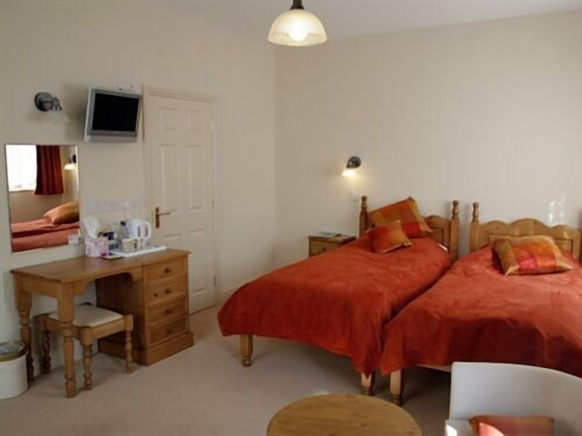 A bedroom with a wooden desk, two beds with red bedding, a television mounted on the wall, and a bedside lamp.