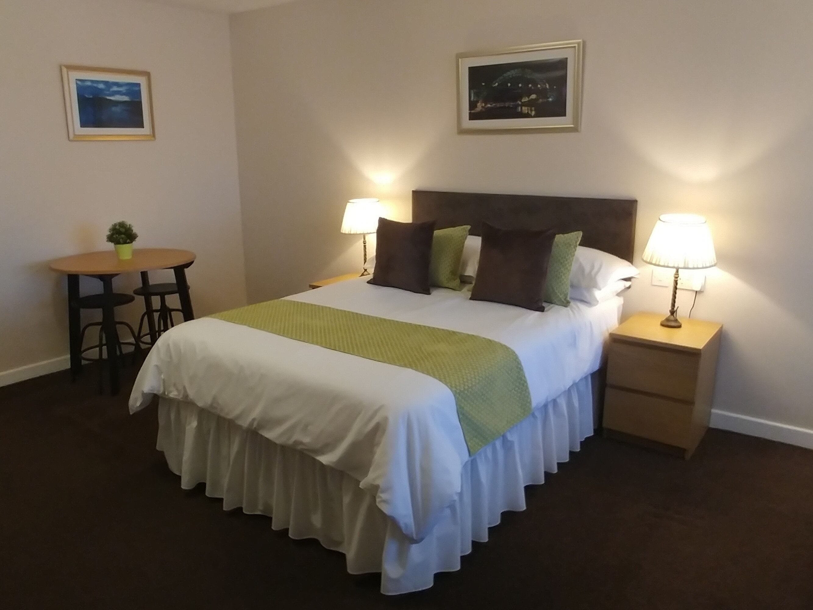 Een netjes opgemaakt bed met een groen-witte spreideken, twee bedlampen, een tafeltje met een plantje en twee ingelijste foto’s aan de muur.