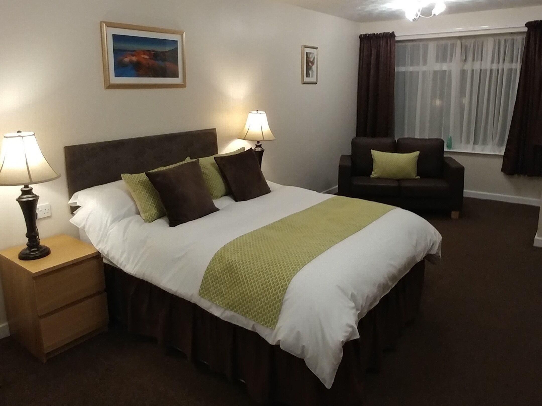 Hotellihuone, jossa on suuri sänky, kaksi yöpöytälamppua, yöpöytä, sohva, ikkuna verhoilla ja seinällä oleva kehystetty taulu.