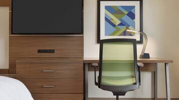 Habitación de hotel con cama, escritorio con computadora, silla, televisor montado en la pared y un cuadro abstracto enmarcado.