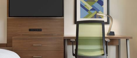 Habitación de hotel con cama, escritorio con computadora, silla, televisor montado en la pared y un cuadro abstracto enmarcado.