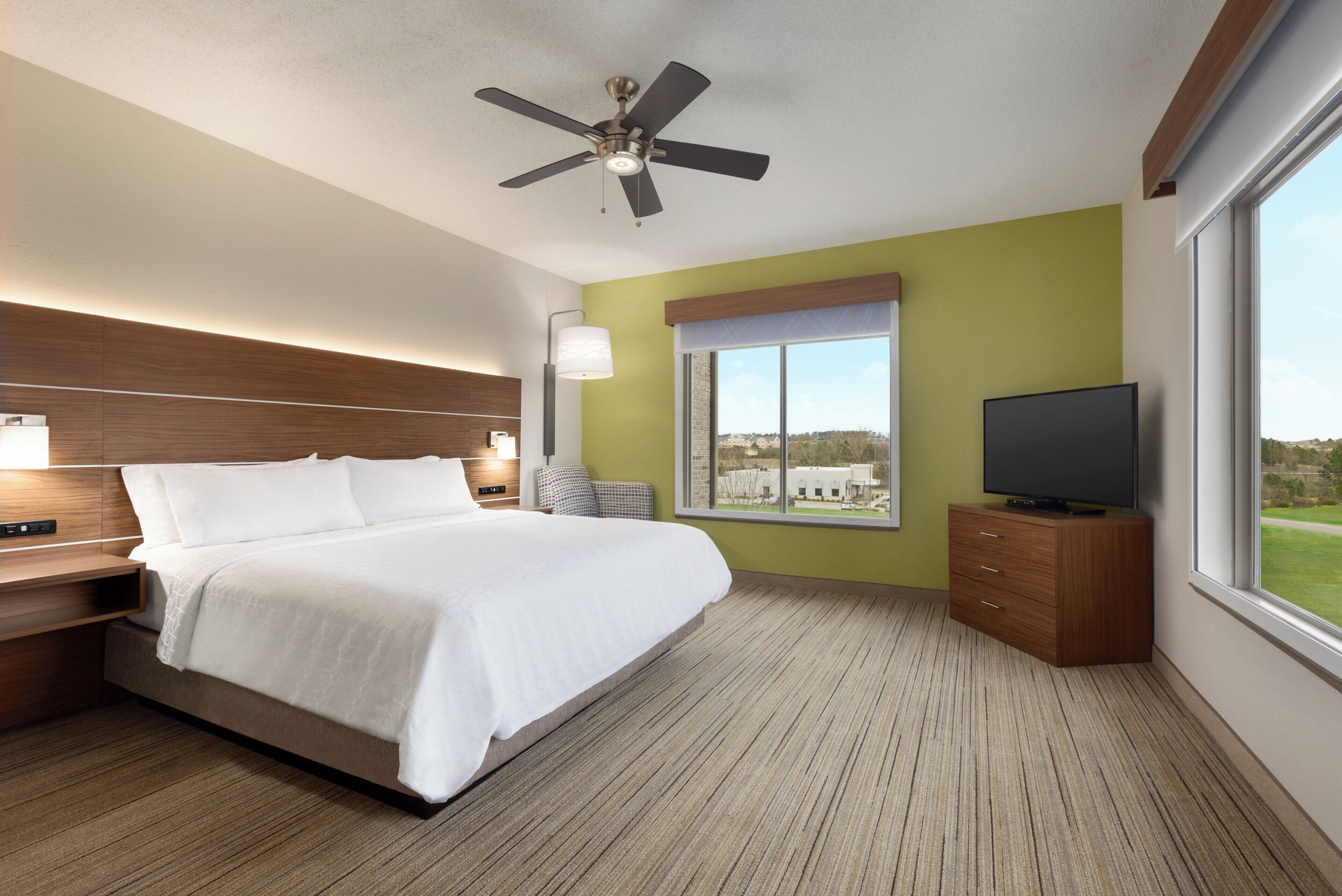 A hotel room with a bed, a television on a wooden cabinet, a ceiling fan, a window with a view, and a bedside table with a lamp.
