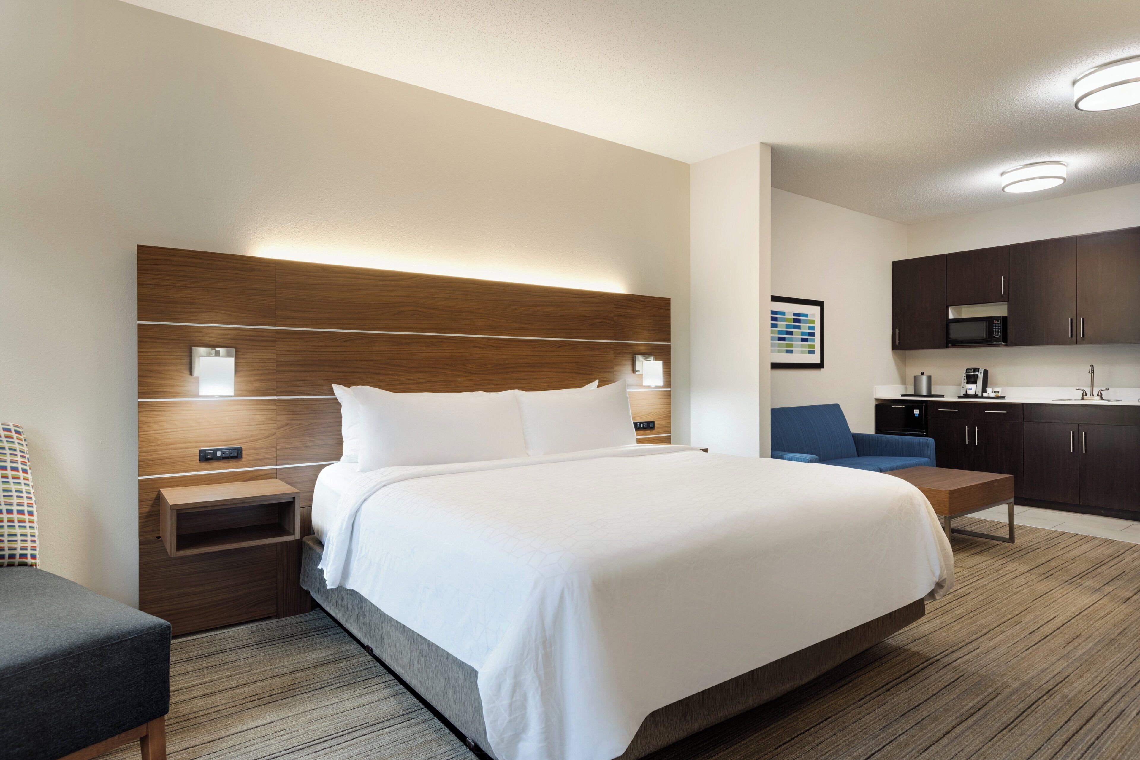 A hotel room with a large bed, a bedside table, a chair, a desk, a kitchenette, and a bathroom.