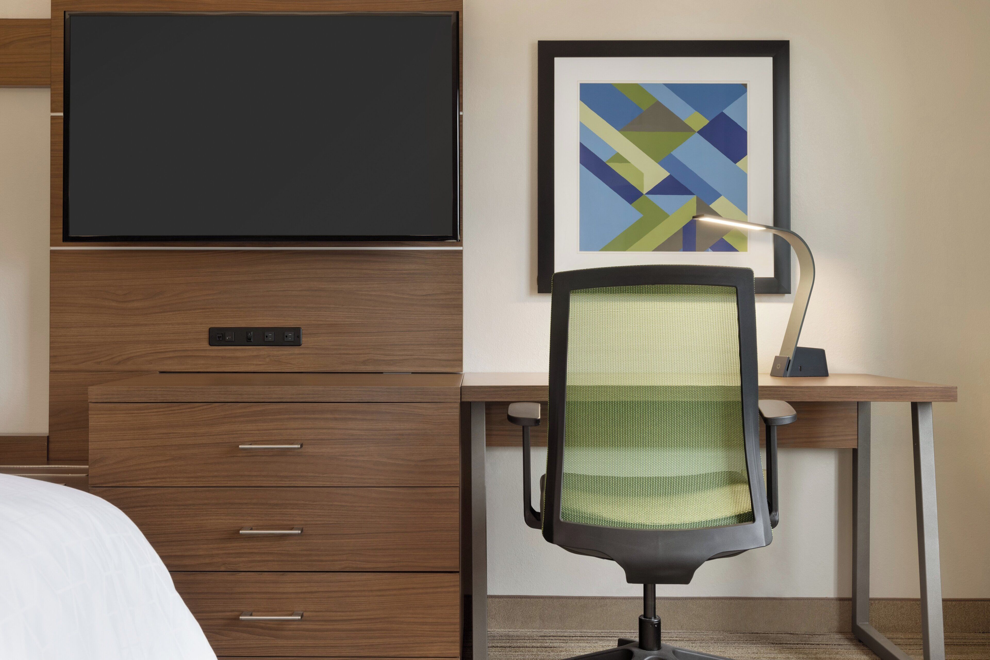 A hotel room with a bed, a desk with a chair, a flat-screen TV, a framed abstract painting, and a power strip on the desk.