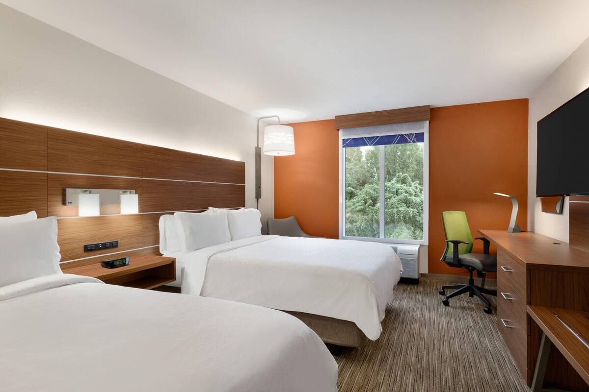 Une chambre d’hôtel avec deux lits, un bureau, une chaise, une télévision et une fenêtre donnant sur des arbres.