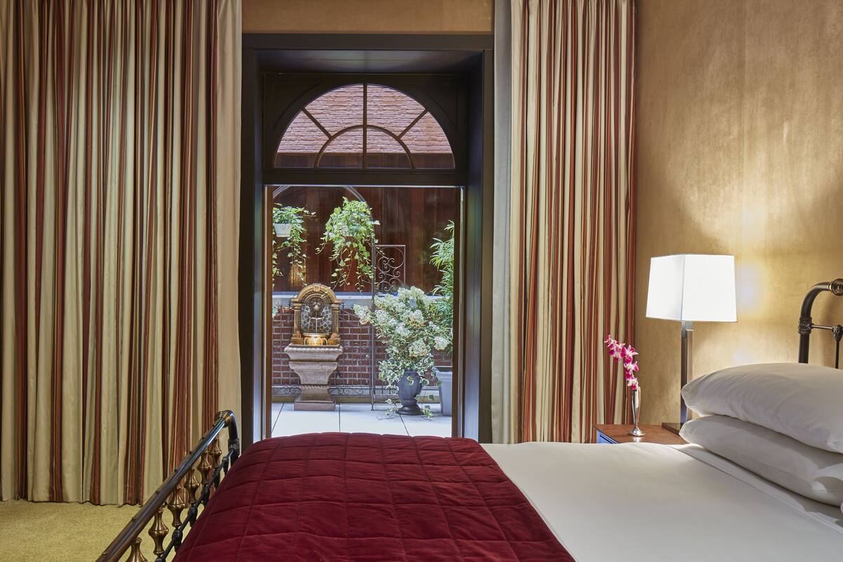 A hotel room with a bed, a nightstand with a lamp, a view of a courtyard with plants and a fountain.