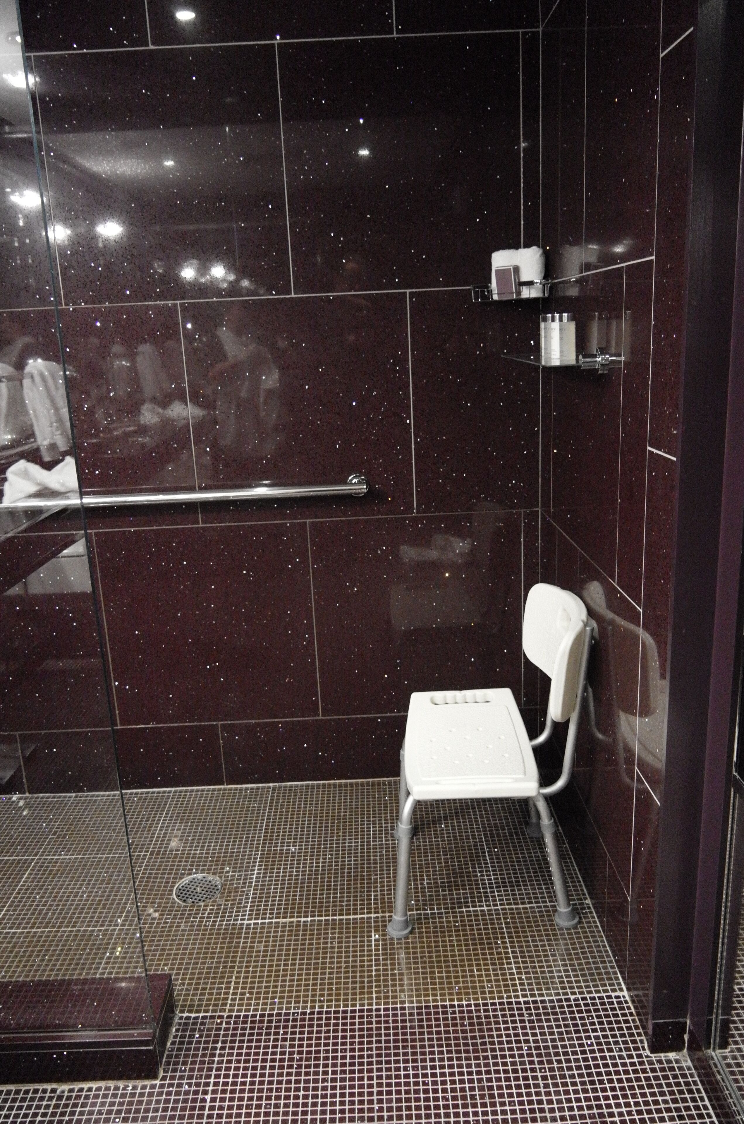 A shower area with a white chair, a handrail, and a tiled floor.