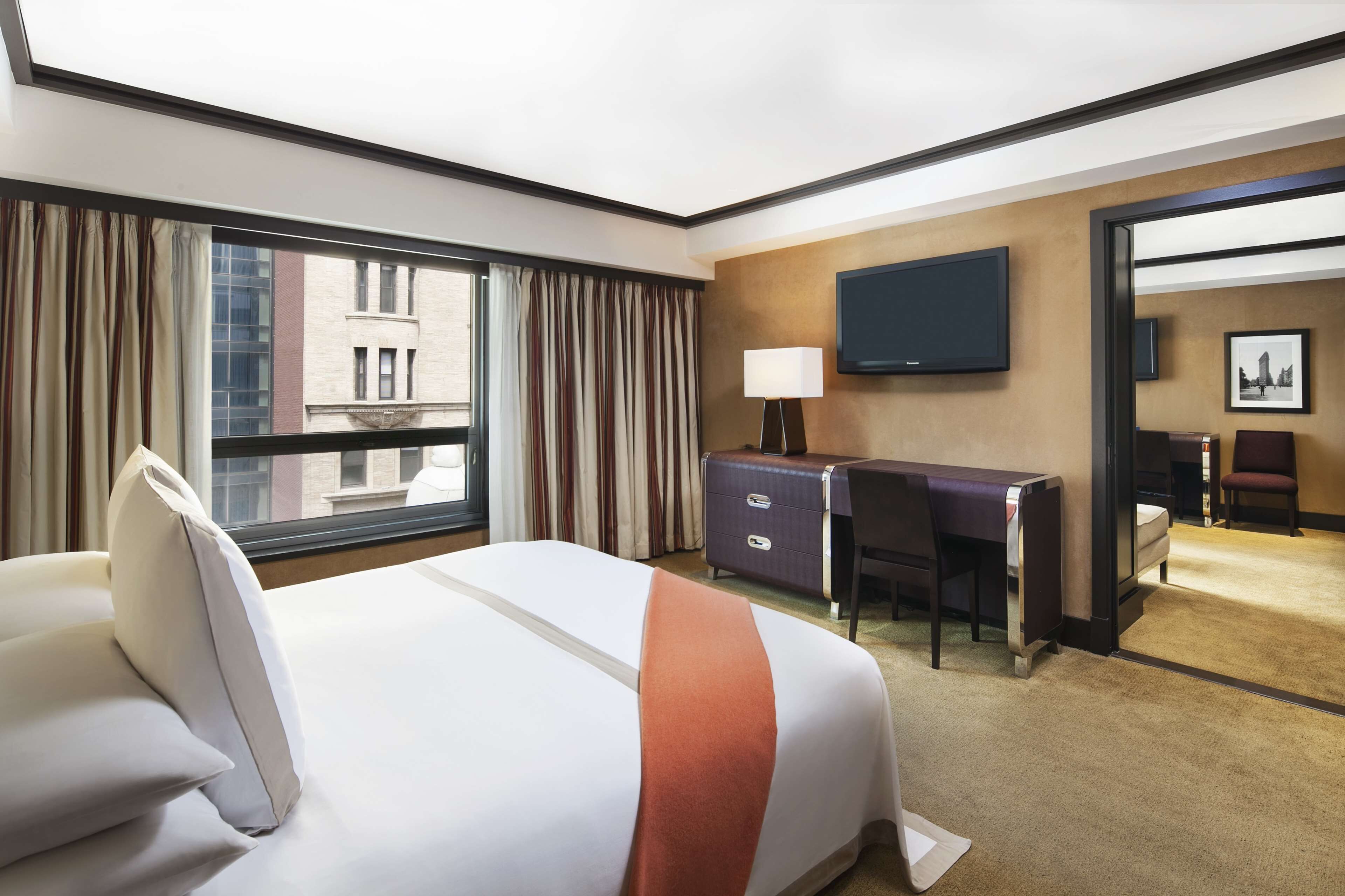 A hotel room with a large bed, a desk, a chair, a TV, and a view of the city through the window.