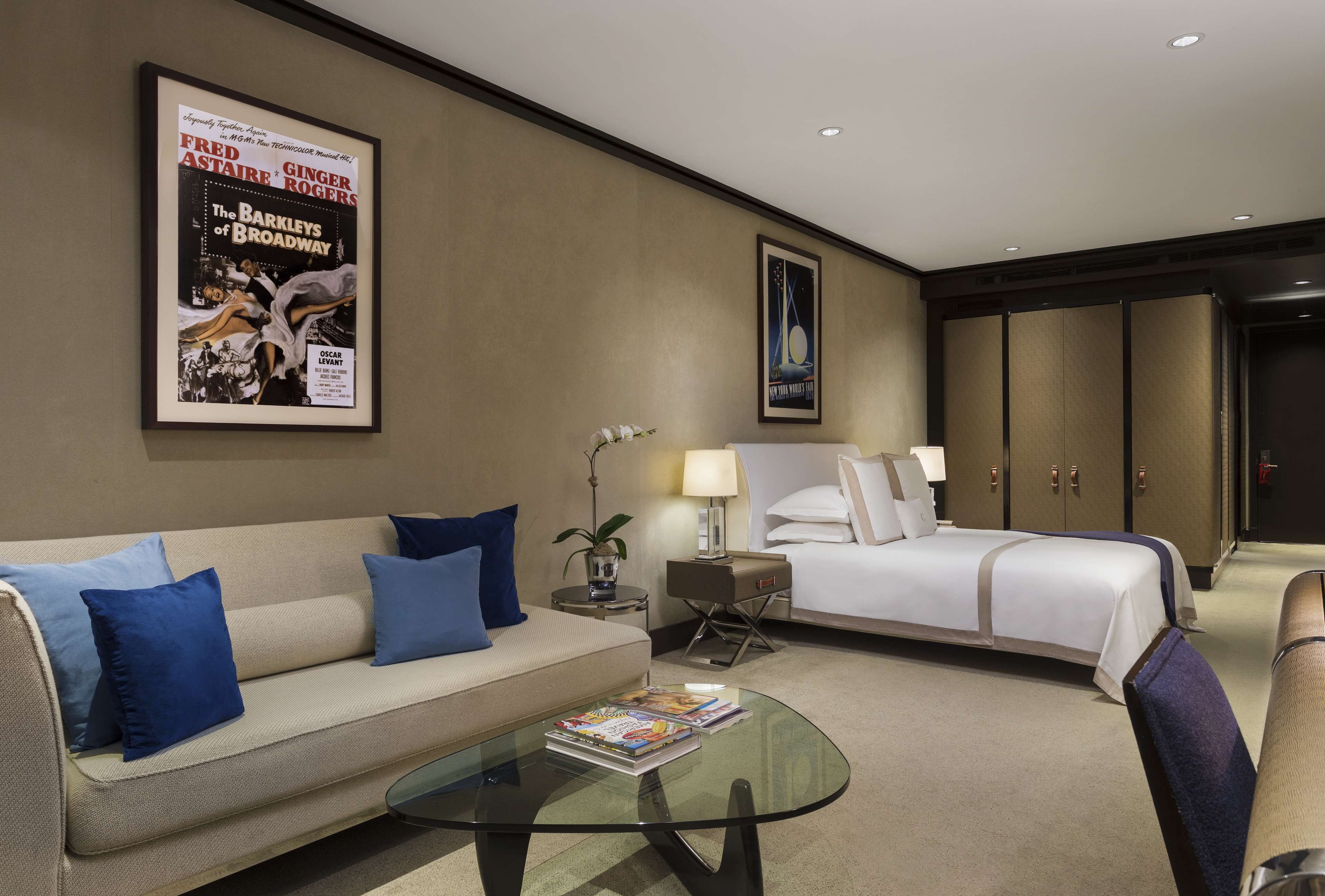 A hotel room with a sofa, a glass coffee table, a bed, a nightstand, and a poster on the wall.