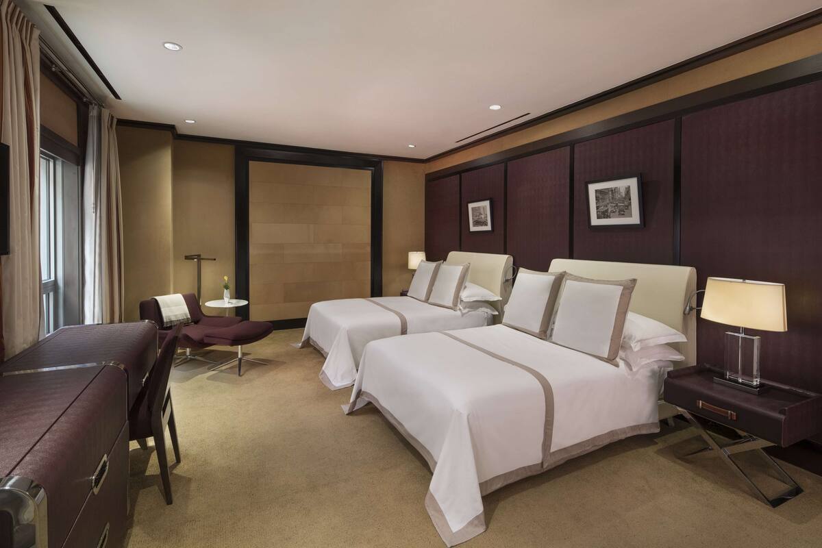 A hotel room with two beds, a desk, a chair, a small table, and a lamp.