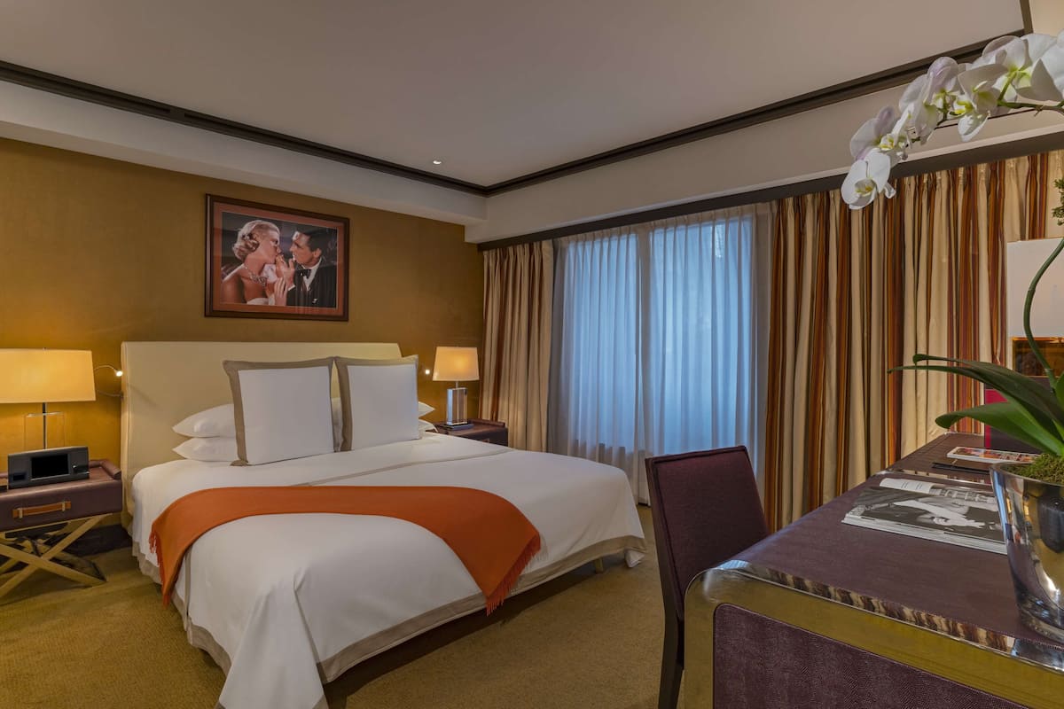 A hotel room with a large bed, two bedside tables with lamps, a desk with a chair, and a large window with curtains.