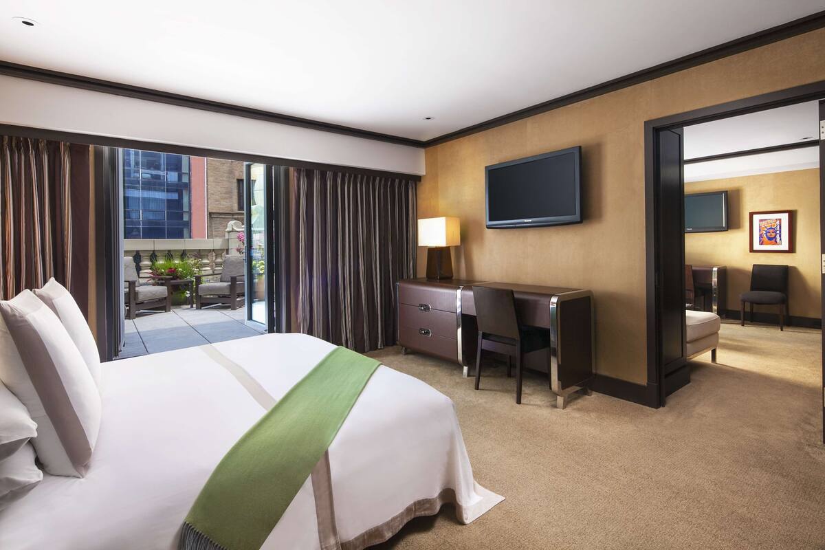 A hotel room with a bed, a desk, a chair, a TV, and a view of the outside.