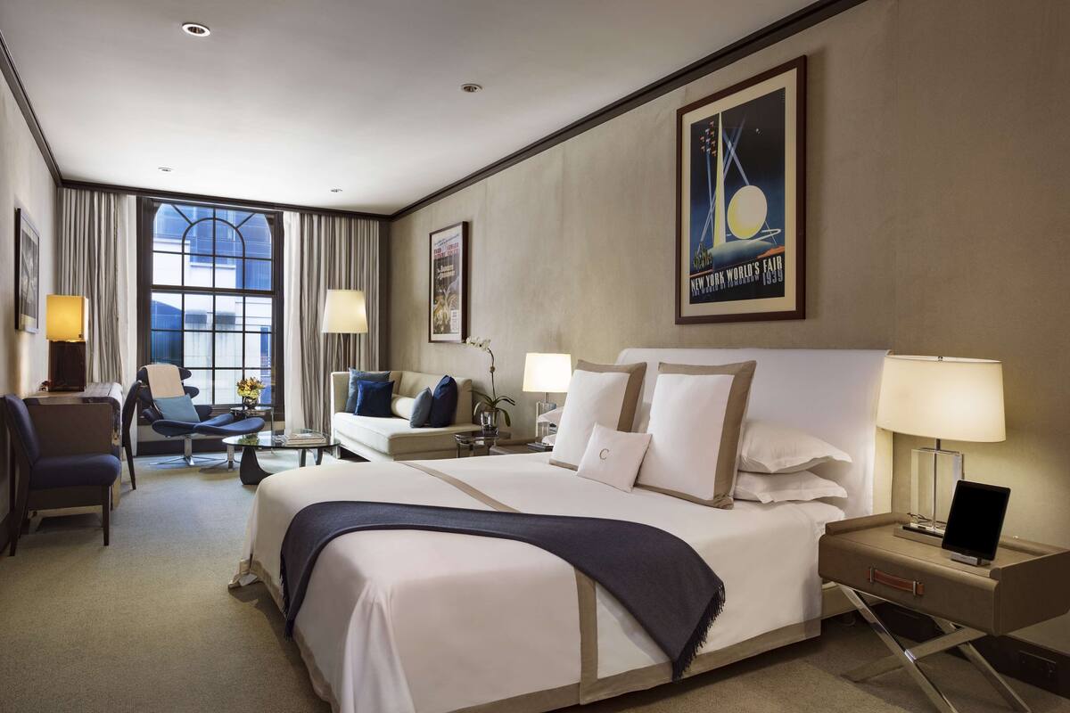 A hotel room with a large bed, a desk, a chair, and a sofa. There is a window with curtains, a lamp, and a framed picture on the wall.