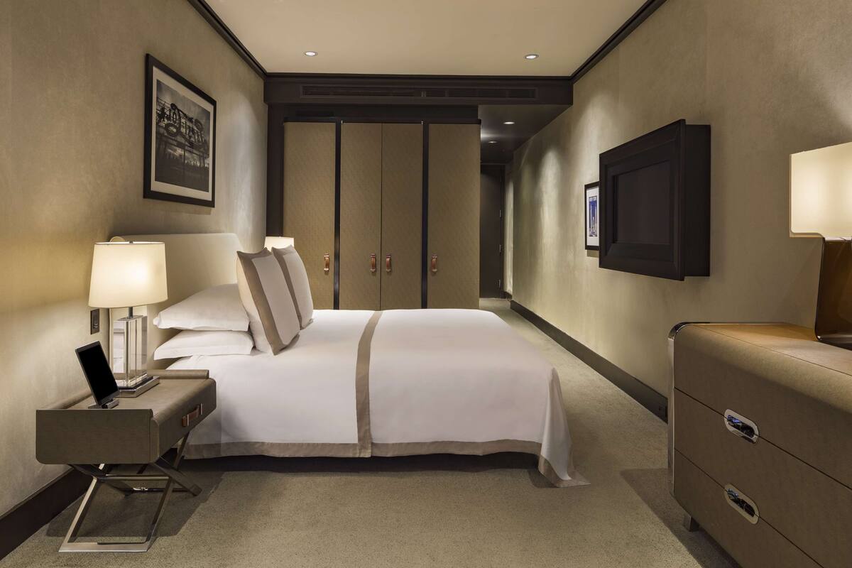 A hotel room with a bed, bedside table with a lamp, a dresser, a television mounted on the wall, and a framed picture on the wall.