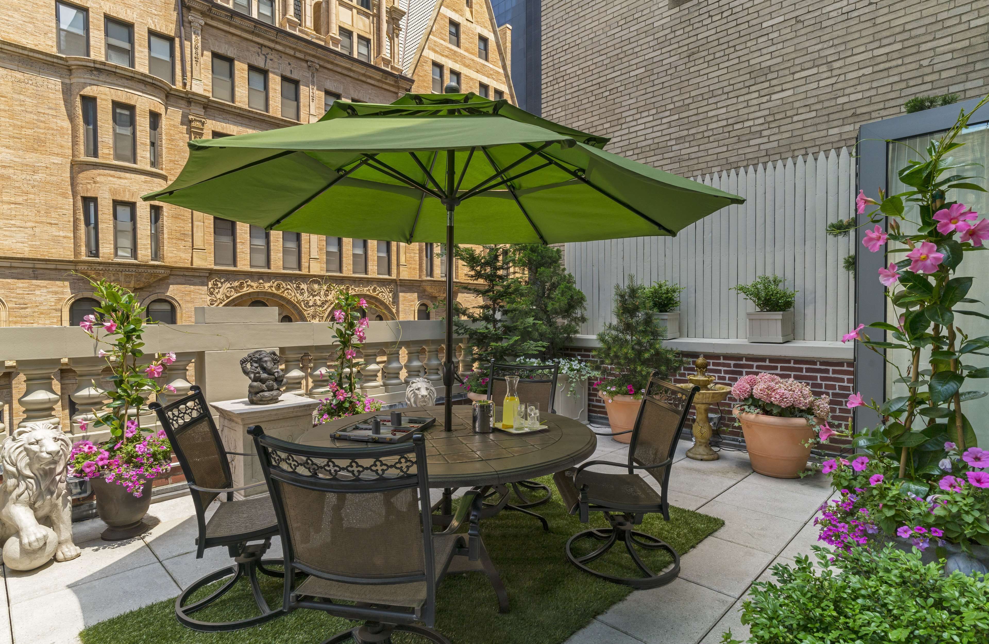 A patio with a round table, chairs, and a green umbrella. There are potted plants and a statue of a lion.