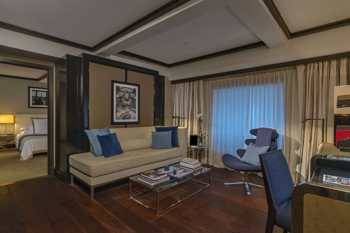 A hotel room with a sofa, armchair, and a coffee table. There is a bed in the background and a window with curtains.