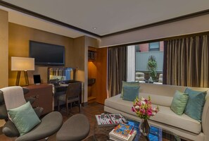 A hotel room with a sofa, armchairs, a desk with a computer, a TV, and a window with curtains.