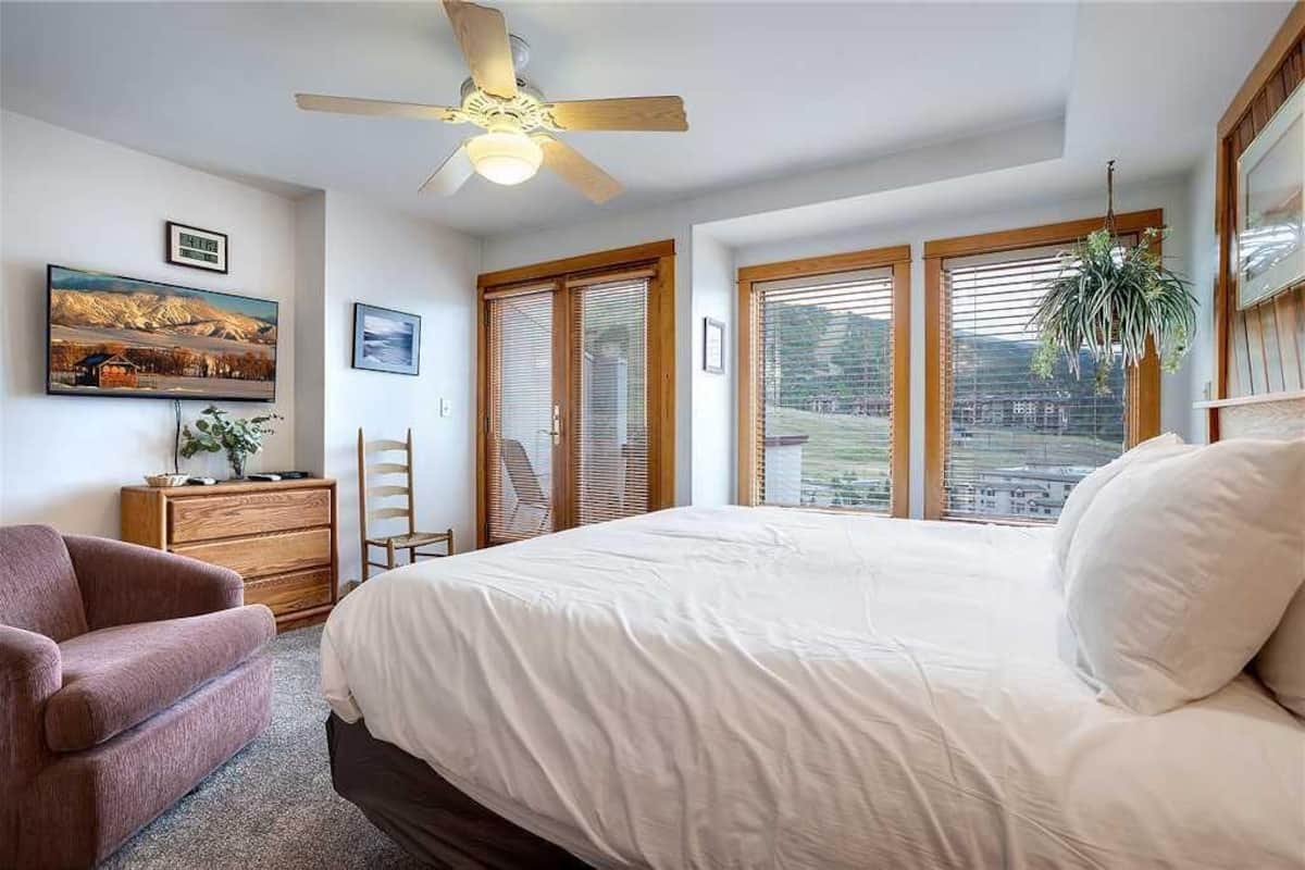 A bedroom with a large bed, a ceiling fan, a dresser, a chair, and a window with blinds.
