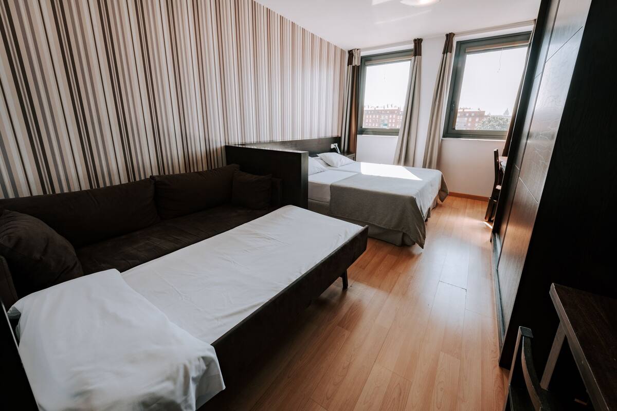 Una habitación de hotel moderna con una cama grande, un sofá y un escritorio. Hay dos ventanas que ofrecen luz natural y vistas a la ciudad.