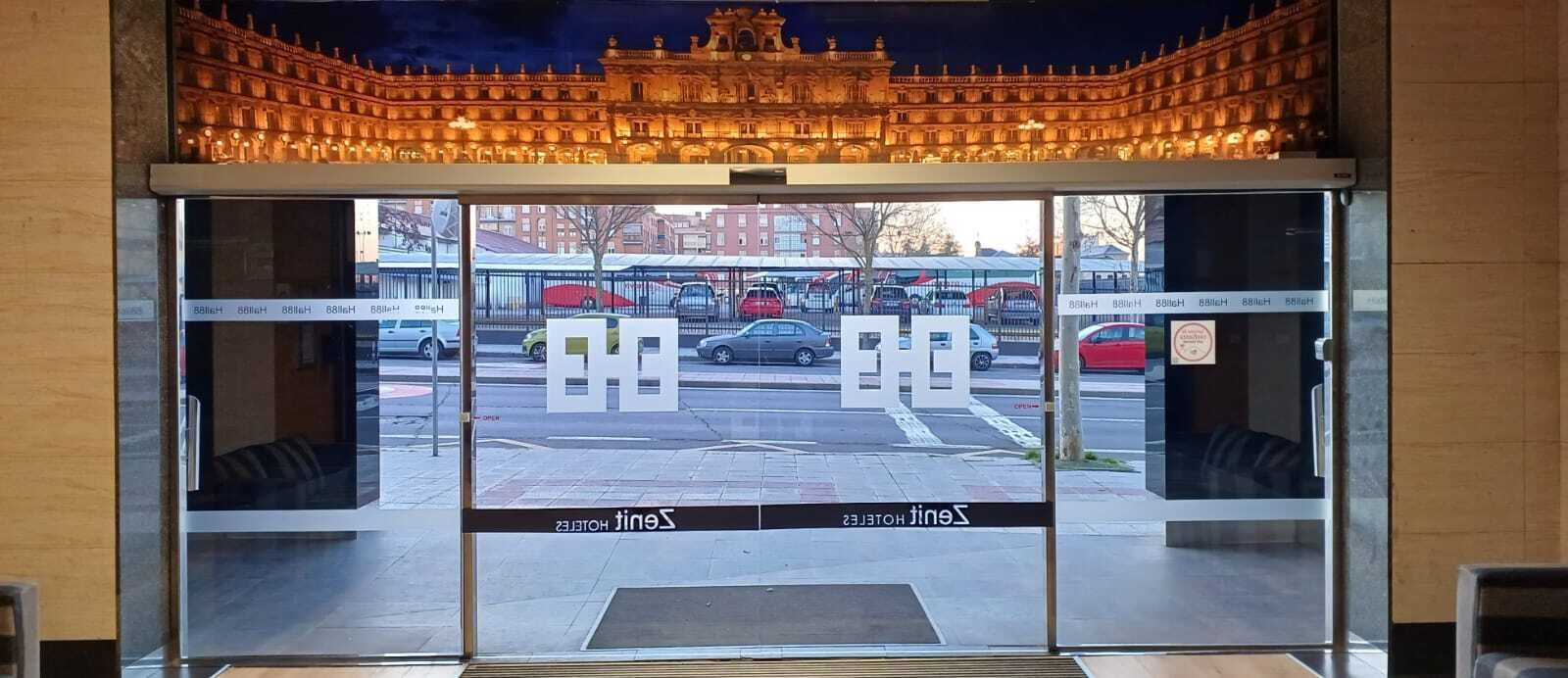 Een hotelentree met een glazen deur, een bord met de naam van het hotel en een zicht op geparkeerde auto's en een gebouw op de achtergrond.