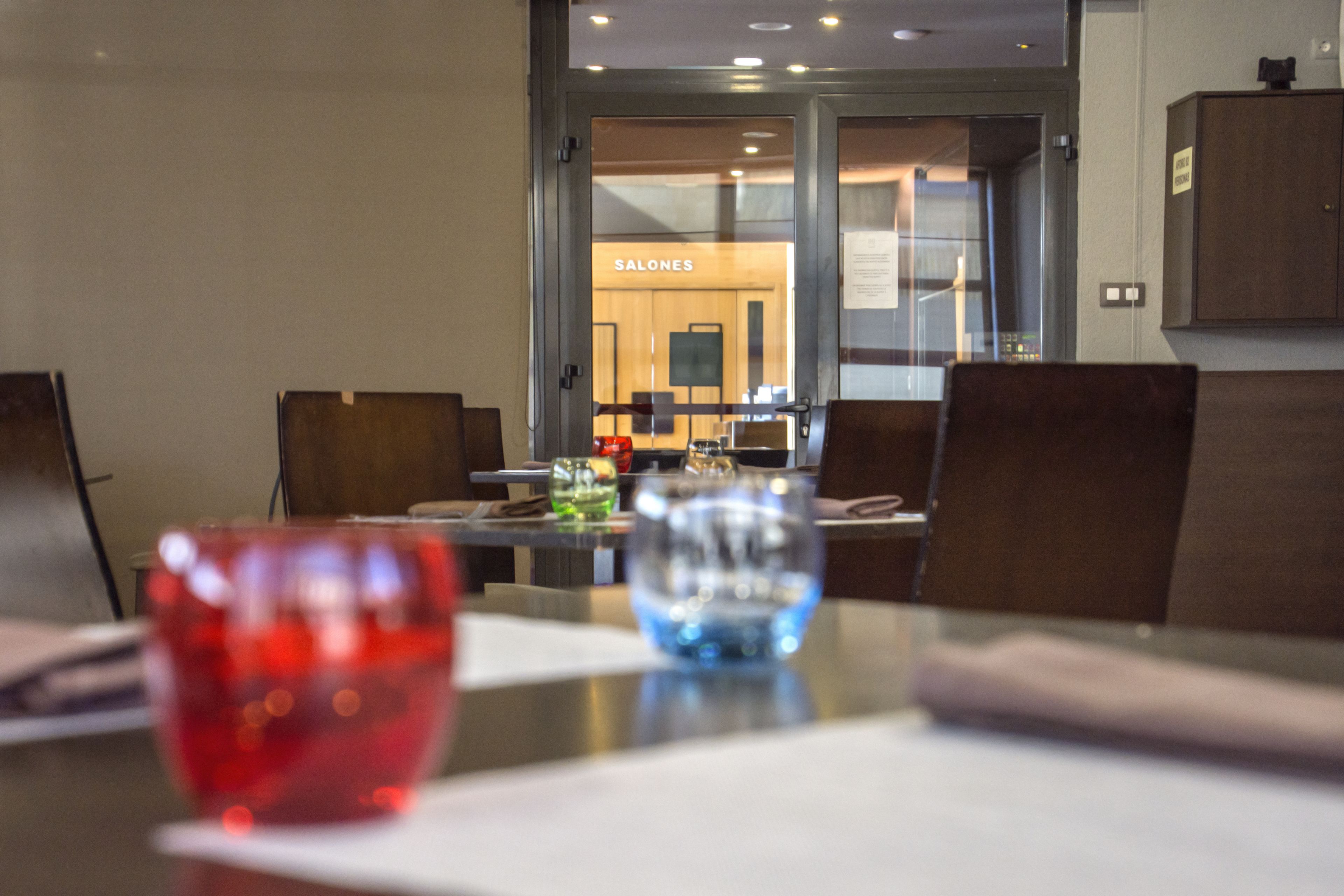 A modern dining area with a glass partition, a red and blue glass on the table, and a sign reading "SALONES" in the background.
