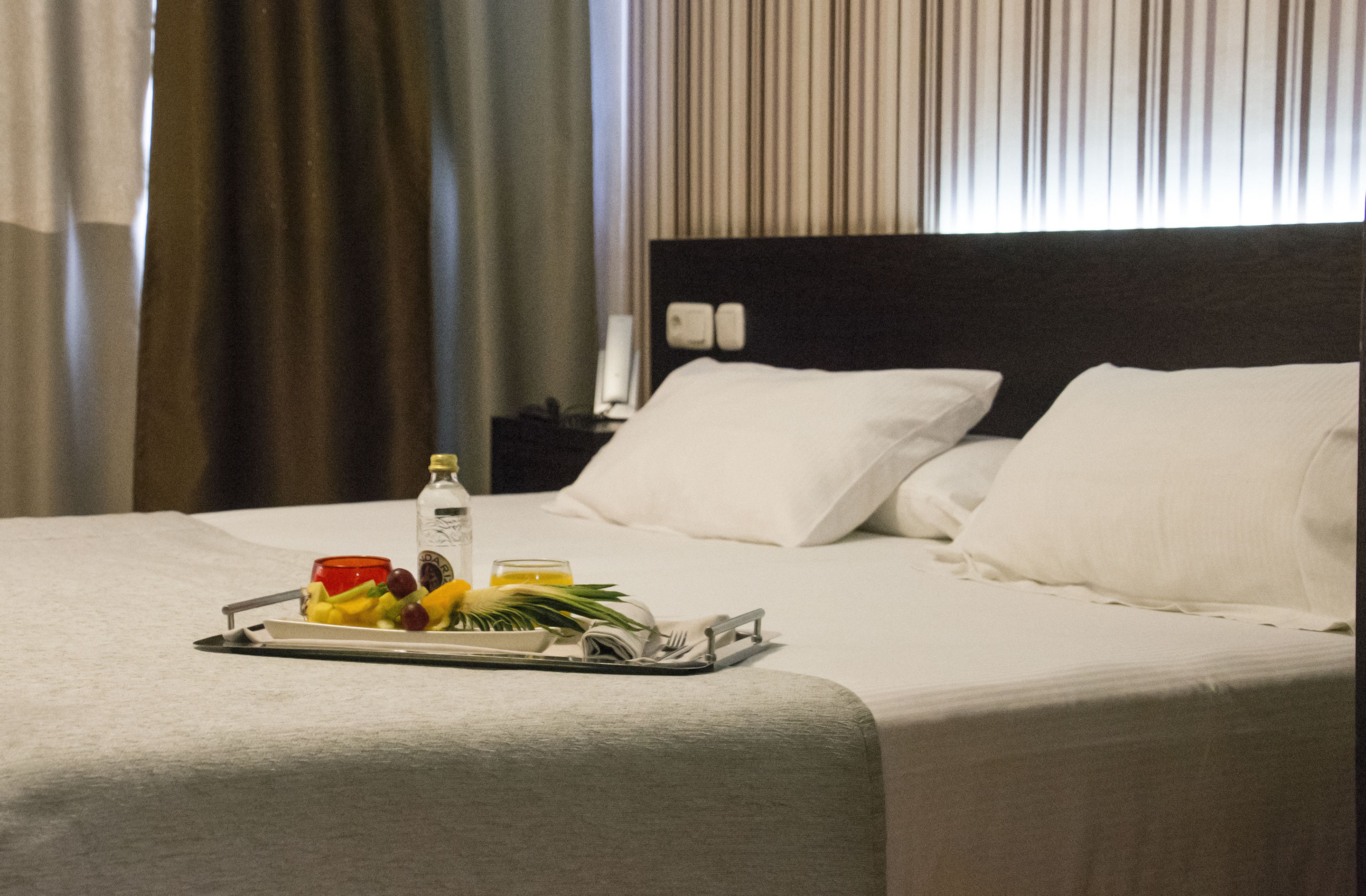 Una cama bien hecha con ropa blanca, una bandeja con frutas y una botella, y una mesita de noche con una lámpara.