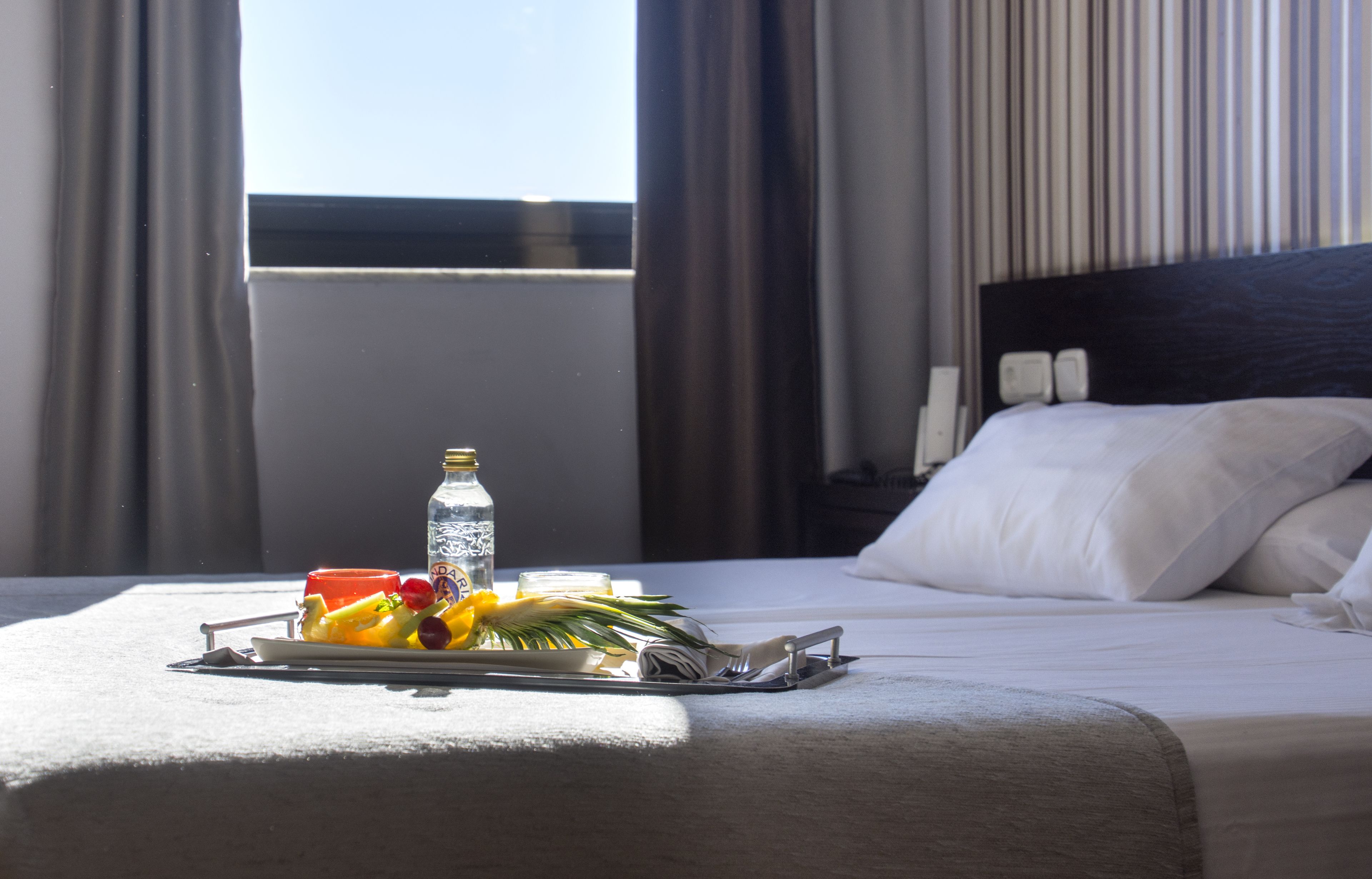 A neatly made bed with white linens, a tray with fruits and a bottle of water, and a nightstand with a lamp.