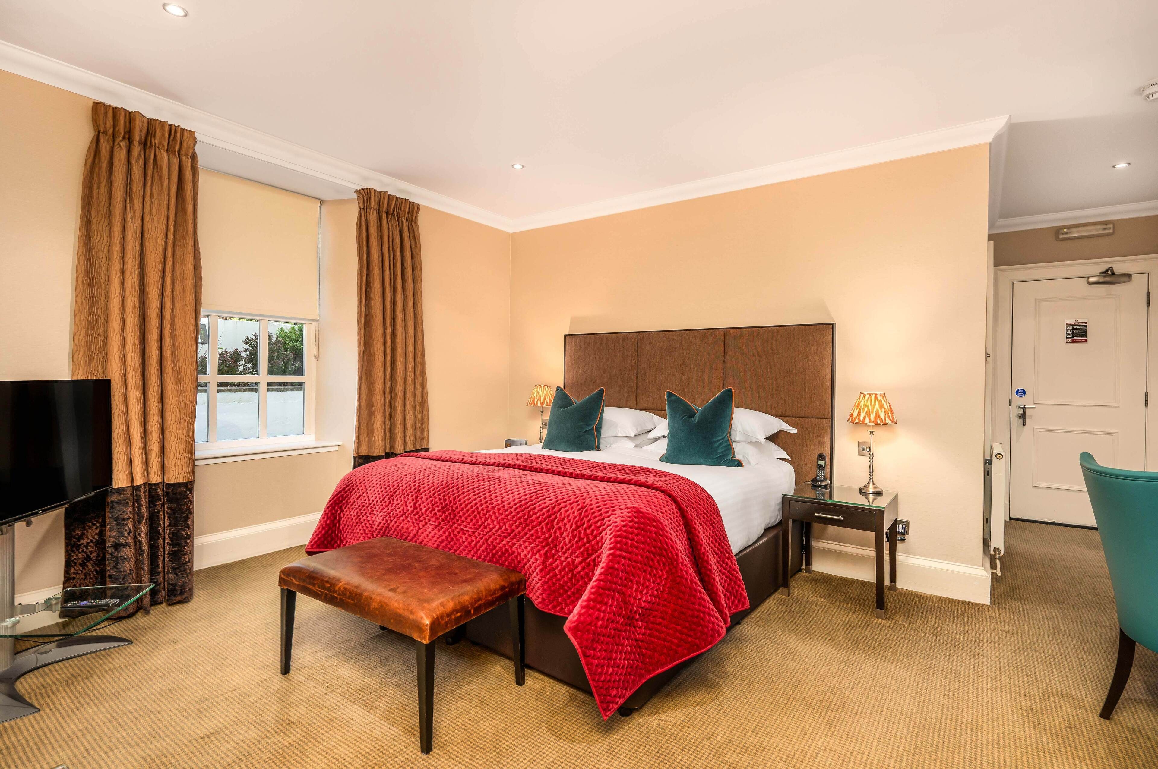 Habitación de hotel con una cama grande, televisión, banco, mesita de noche con lámpara y una puerta.