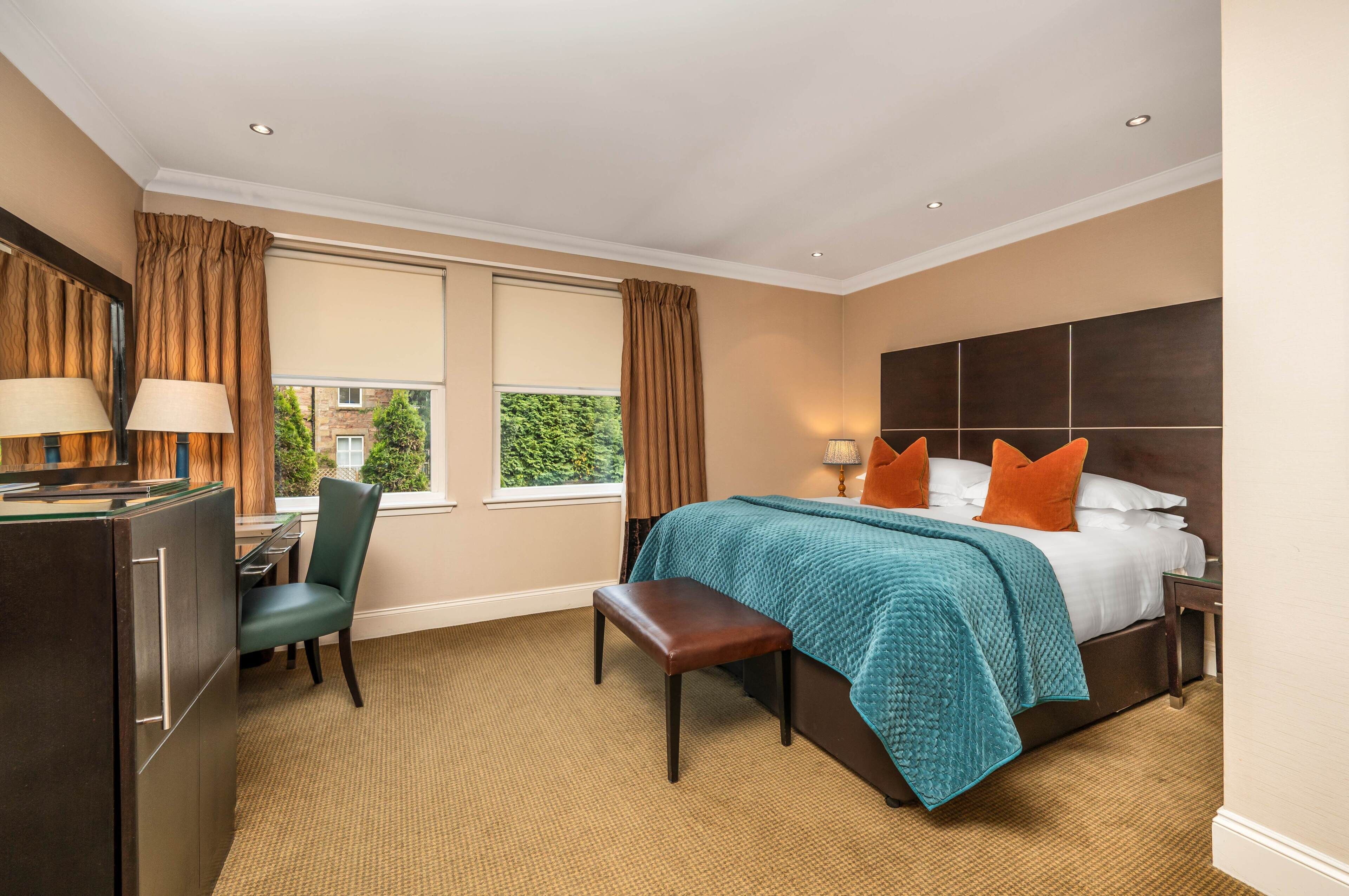 A hotel room with a bed, a desk, a chair, a bench, a window with curtains, and a view of greenery outside.