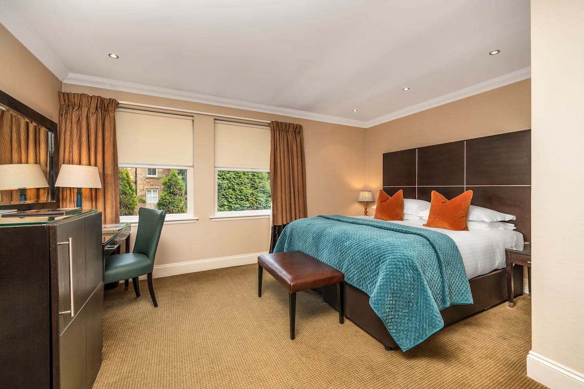 A hotel room with a bed, a desk, a chair, a bench, a dresser, and a window with curtains.