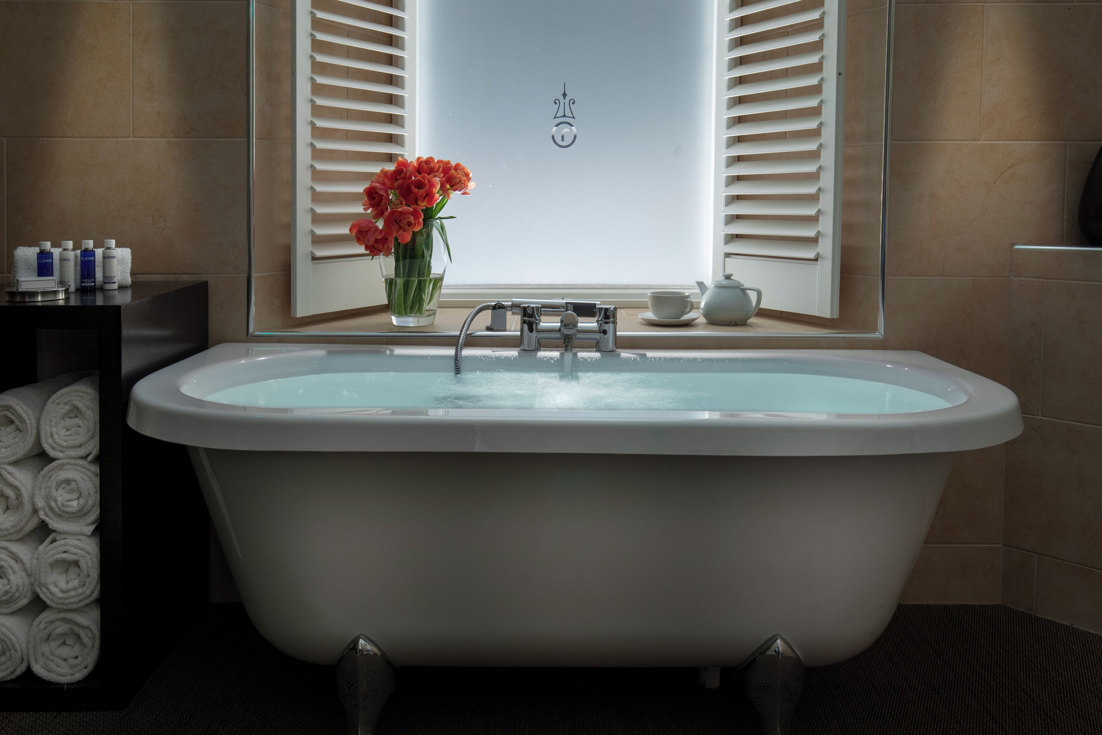 Une salle de bain avec une baignoire sur pieds, un miroir avec stores, un vase de fleurs et des articles de toilette rangés sur une étagère.